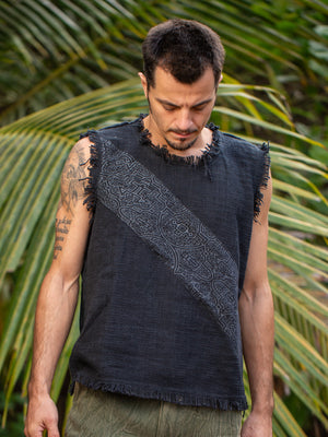 A man wearing the Shipibo Tank Top—sleeveless and textured in black with a patterned diagonal stripe—stands outdoors beside green palm leaves, looking down.