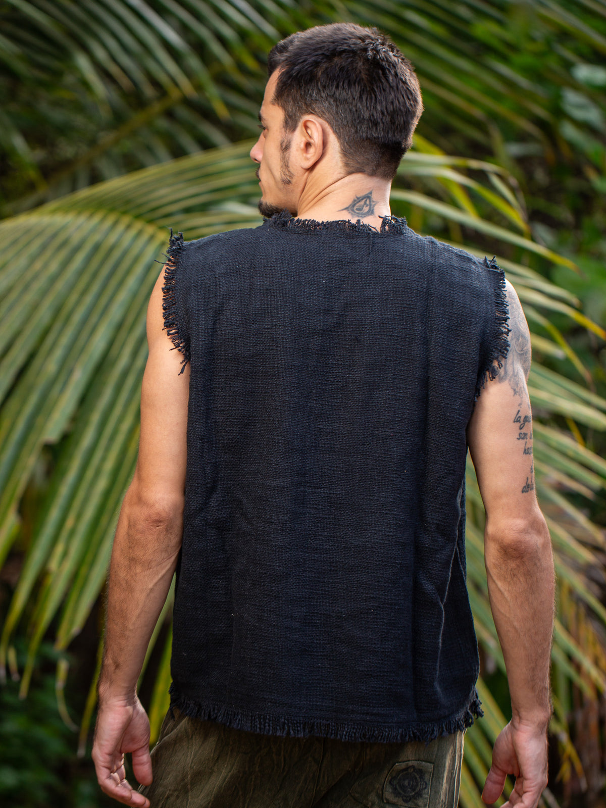 A man stands outdoors with his back to the camera, wearing the Shipibo Tank Top—a sleeveless black textured piece. Palm leaves frame the background, while tattoos are visible on his right arm and neck.