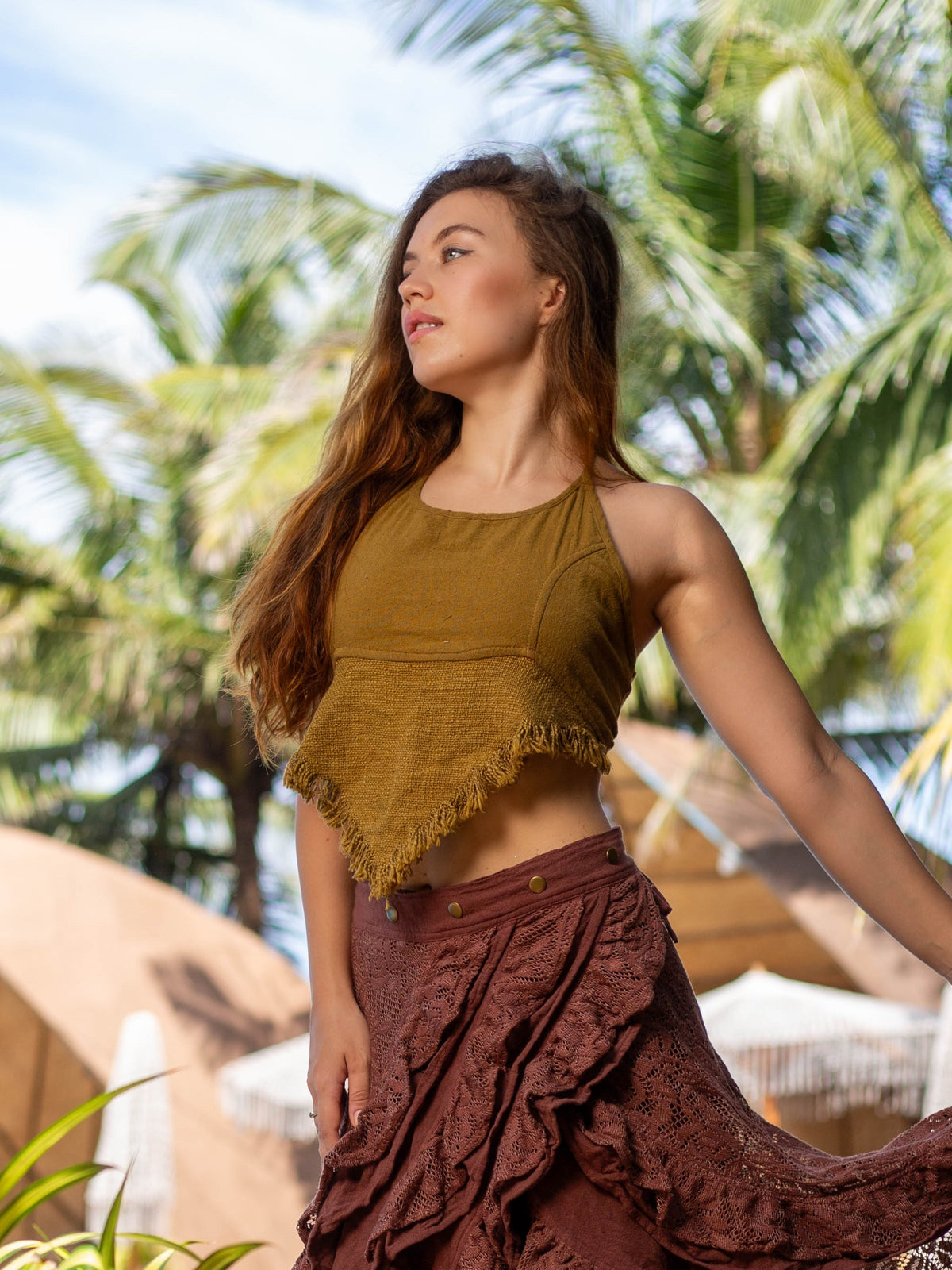A woman with long hair gazes aside outdoors, wearing a Shanti Crop Top with fringe paired with a flowing skirt. The boho gypsy design harmonizes with the palm trees and clear sky, creating a tropical ambiance.