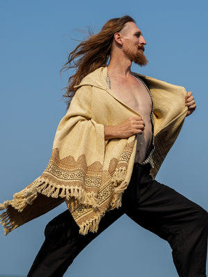 A person with long hair and a beard poses outdoors against a clear blue sky, wearing the Swing Shawl—a patterned beige blockprint shawl with fringes—paired with black pants.