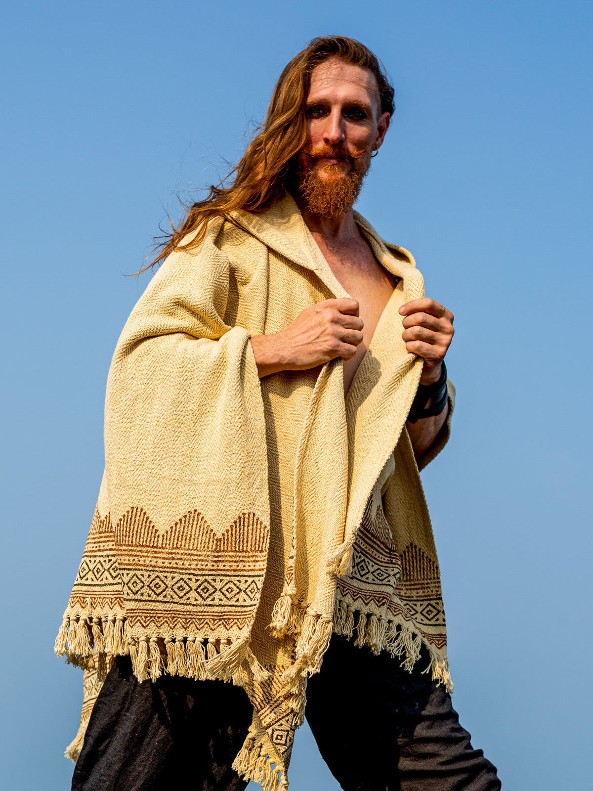 A person with long red hair and a beard wears the Swing Shawl in tan blockprint and dark pants, posing confidently outdoors against a clear blue sky.