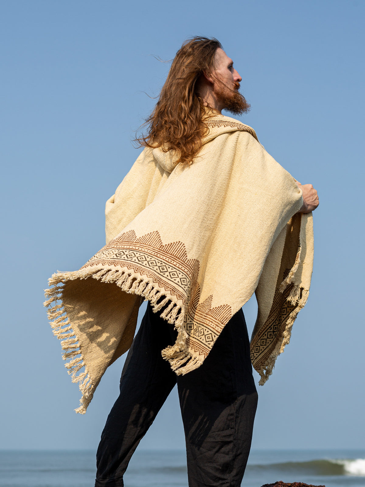 A person with long hair stands outdoors, draped in the Swing Shawl—a beige blockprint wrap featuring fringed edges and geometric patterns—facing sideways against a clear blue sky.
