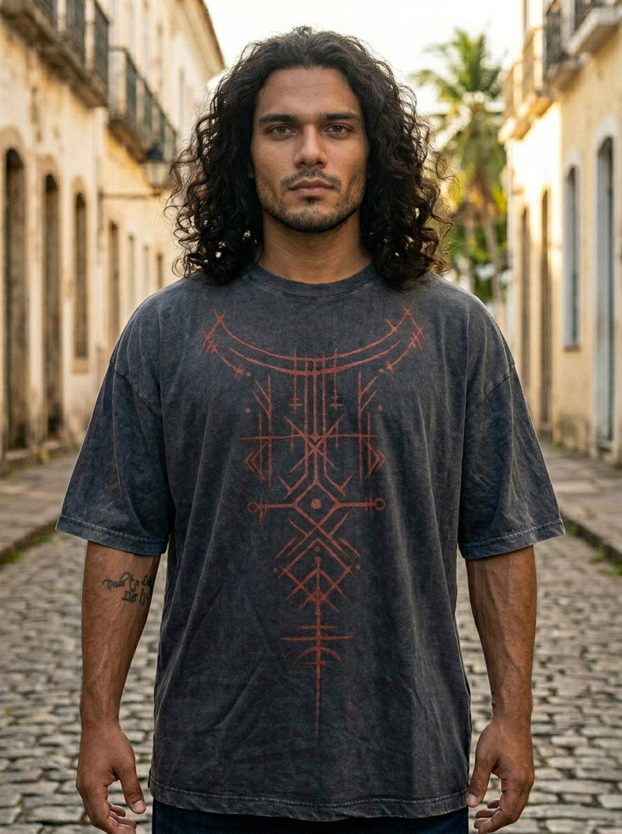 A man with long curly hair stands on a cobblestone street between old buildings, wearing the Rune Ember Tee—an oversized shirt featuring a geometric red pattern in classic streetwear style—as he looks directly at the camera.