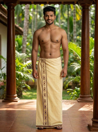 A smiling man stands barefoot on a tiled floor, wearing the Mehfil Dhoti—a traditional cream-colored cotton dhoti with a decorative border that adds an ethnic touch. He is shirtless, accessorized with a necklace, and surrounded by lush green plants and trees outdoors.