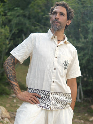 A man with a patterned tattoo on his right arm stands outdoors, thoughtfully wearing the Vairagya Shirt—crafted from light, organic cotton and distinguished by its unique geometric designs on the pocket and waist. His conscious fashion choice is complemented by a backdrop of lush trees and greenery.