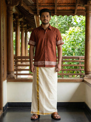 A man stands on a veranda wearing the Adhir Shirt, an ethnic contemporary brown short-sleeve cotton shirt with patterns, paired with a traditional white mundu featuring a yellow border and brown sandals. Lush greenery is visible in the background.