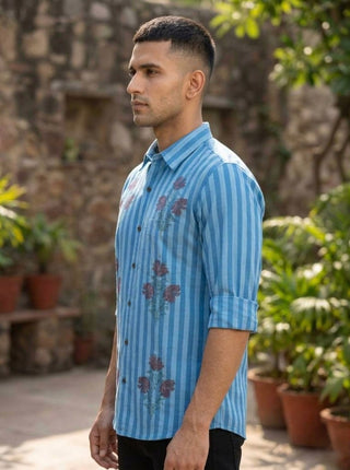 A man with short hair stands sideways outdoors, wearing the Neelpushpa Shirt—sky blue with delicate floral stripes. Potted plants and a stone wall are visible in the background.