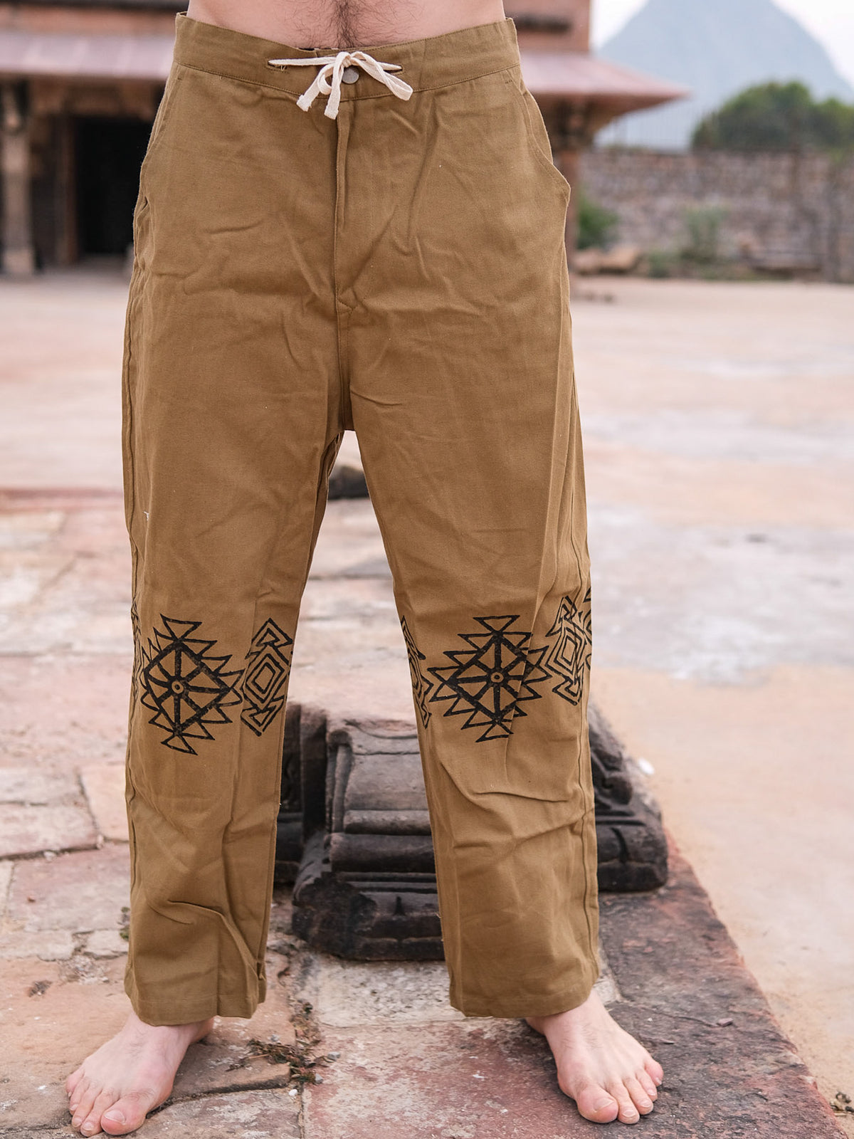 A person stands barefoot on stone ground, wearing Tribal Pants made from 100% cotton with hand-blockprinted black geometric patterns on the thighs. The drawstring waistband is visible, and an outdoor structure appears in the background.