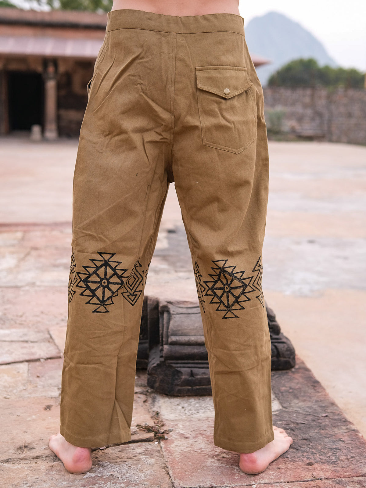 Barefoot outdoors, a person faces away wearing Tribal pants—100% cotton with black geometric hand-block prints on the back of the legs and a buttoned back pocket. Stone structures and distant mountains can be seen behind.