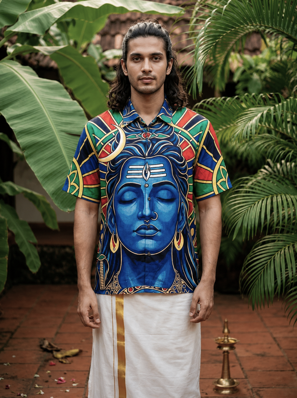 A man stands outdoors surrounded by tropical plants and a brass lamp on a tiled floor, wearing the blue Aadi Dev Shirt featuring a vibrant Shiva-inspired design along with a traditional white dhoti.