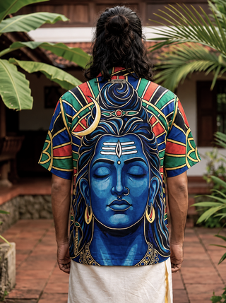 Wearing the Aadi Dev Shirt, a person with long hair is seen from behind, outdoors among tropical plants. The shirt is inspired by Shiva and features a vibrant, detailed painting of Lord Shiva’s blue face surrounded by colorful patterns.