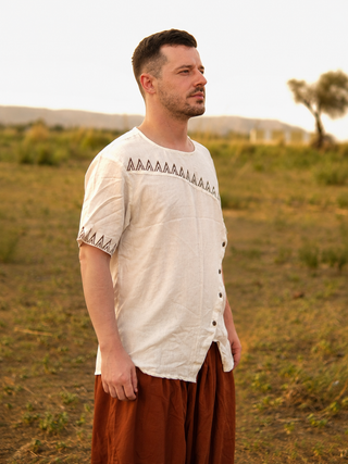 A man with short hair and a trimmed beard stands outdoors on a grassy field, wearing the light-colored Origin Kurta with decorative trim and loose brown pants, with hills and a tree in the background.