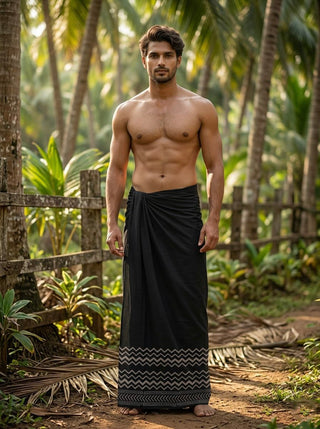 A shirtless man stands barefoot on a dirt path surrounded by tropical plants and palm trees, wearing the Midnight Zigzag Dhoti with its distinctive black fabric and zigzag border.