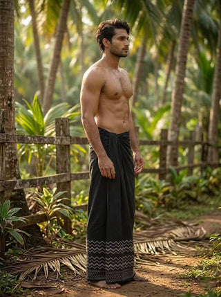 A shirtless man stands outdoors on a dirt path, surrounded by lush green palm trees and a wooden fence in the background, wearing the Midnight Zigzag Dhoti—a traditional black dhoti featuring a subtle zigzag border.