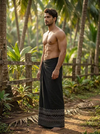 A shirtless man stands on a dirt path in a tropical setting, wearing the Midnight Zigzag Dhoti with its distinctive zigzag border near the hem. Palm trees and lush greenery surround him as sunlight filters through the leaves.