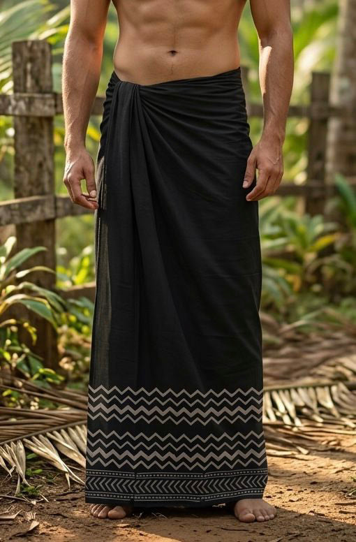 A shirtless man stands outdoors, surrounded by tropical plants and a wooden fence, wearing the Midnight Zigzag Dhoti—a black wrap featuring striking zigzag and geometric patterns near the bottom.