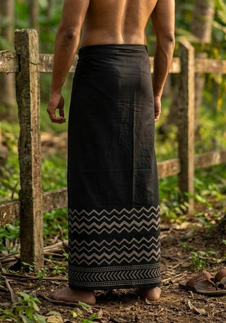 A barefoot person stands outdoors on a dirt path, wearing the Midnight Zigzag Dhoti with a zigzag border near the bottom. Their back faces the camera, and a wooden fence and greenery can be seen in the background.