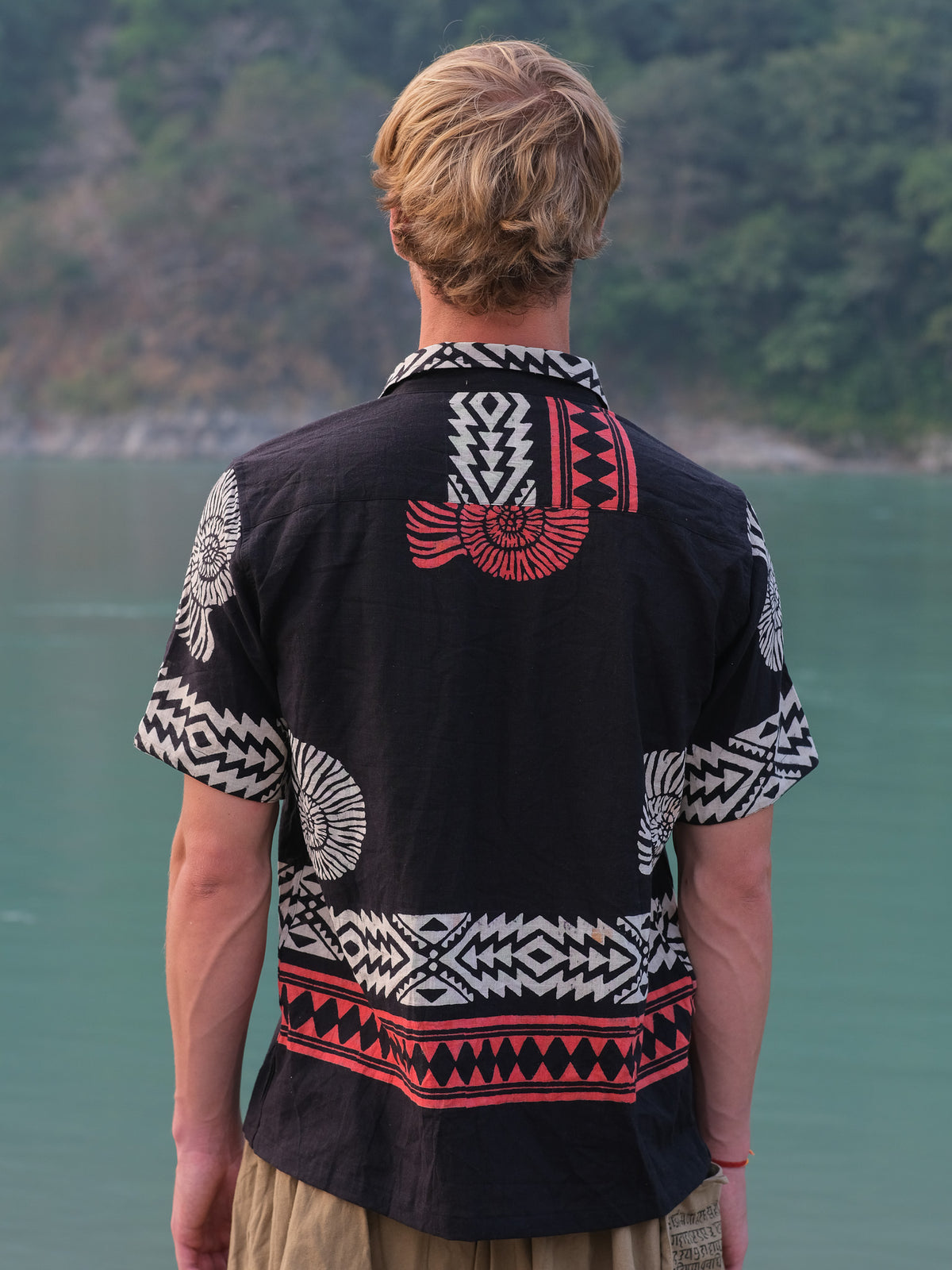 A person with blond hair, seen from behind, wears the Belgium Shirt featuring bold white and red geometric patterns, standing near a body of water with trees and hills in the background.