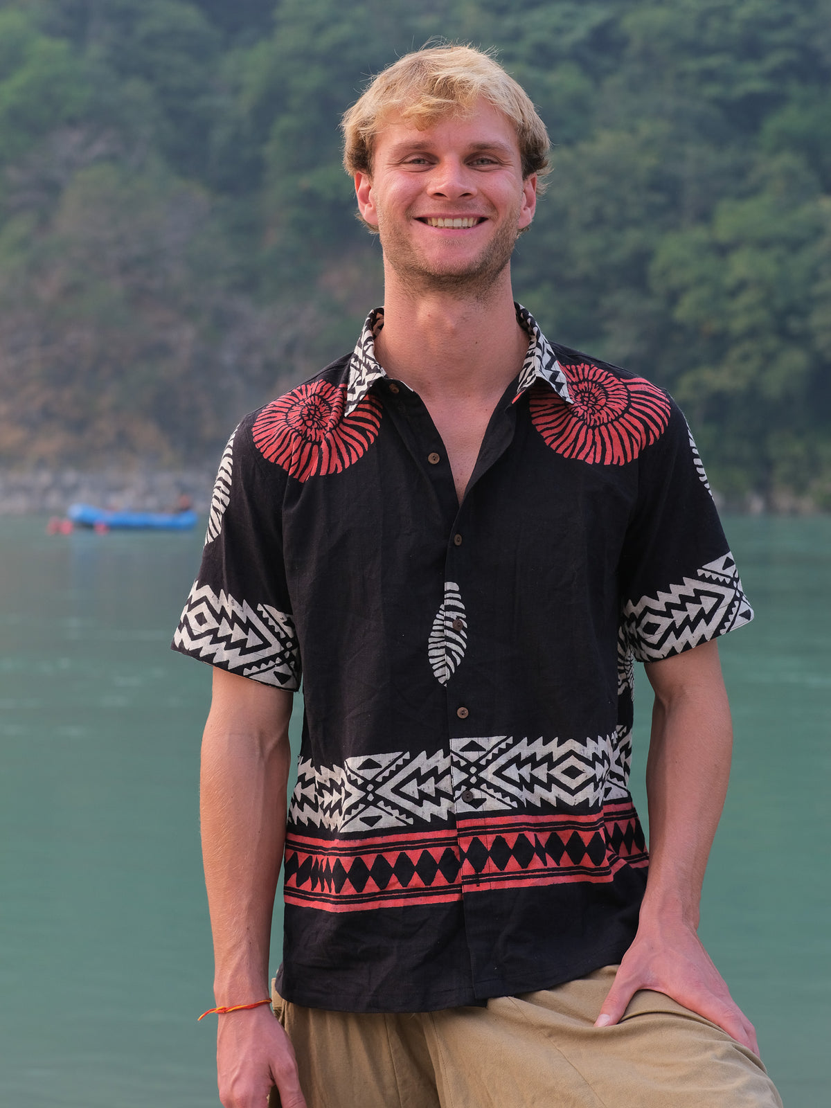 A young man with blond hair, wearing the Belgium Shirt—featuring a black and red bohemian pattern—and beige pants, smiles while standing beside a green river with a forested background.
