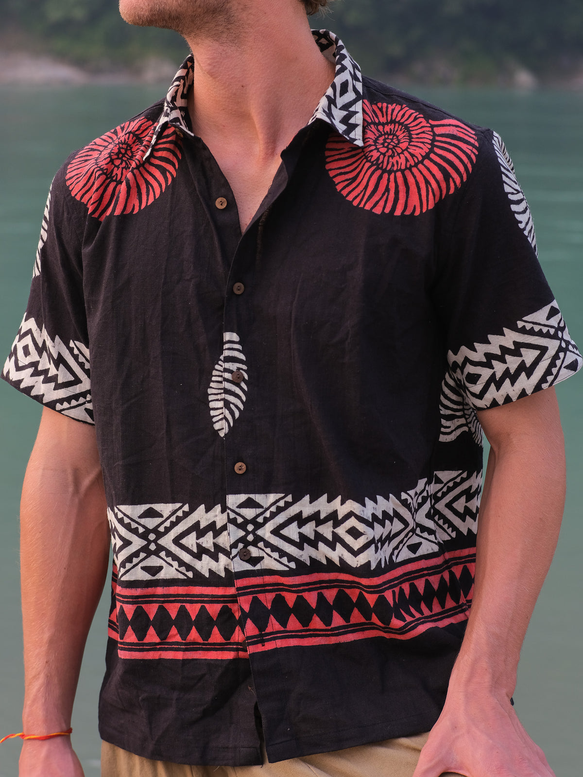 A man stands near water wearing the Belgium Shirt, a black short-sleeve top with bold red and white geometric and shell patterns inspired by Rajasthani handblock prints; his face is partially out of frame for a bohemian vibe.