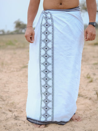 Standing barefoot on sandy ground, a person wears a Kangura Dhoti with a Rajasthani handblock print border and a black wristwatch on their right wrist, while their upper body remains hidden.