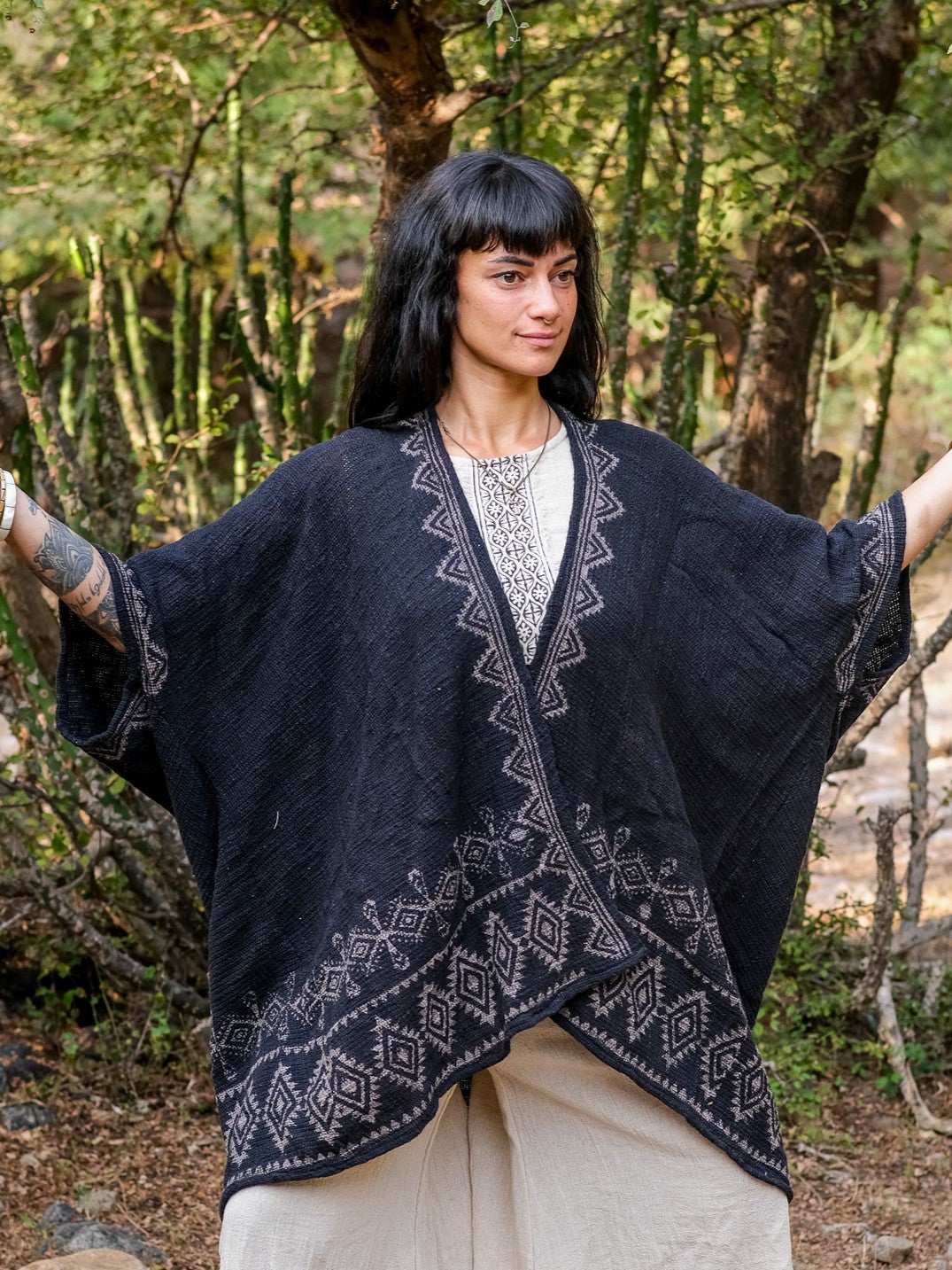 A woman with dark hair and bangs stands outdoors among lush greenery, arms slightly raised, wearing the Miraya Shrug over a light dress.