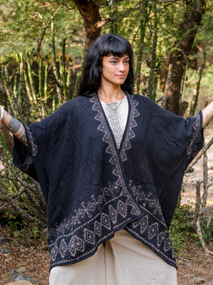 A woman with dark hair and bangs stands outdoors among lush greenery, arms slightly raised, wearing the Miraya Shrug over a light dress.