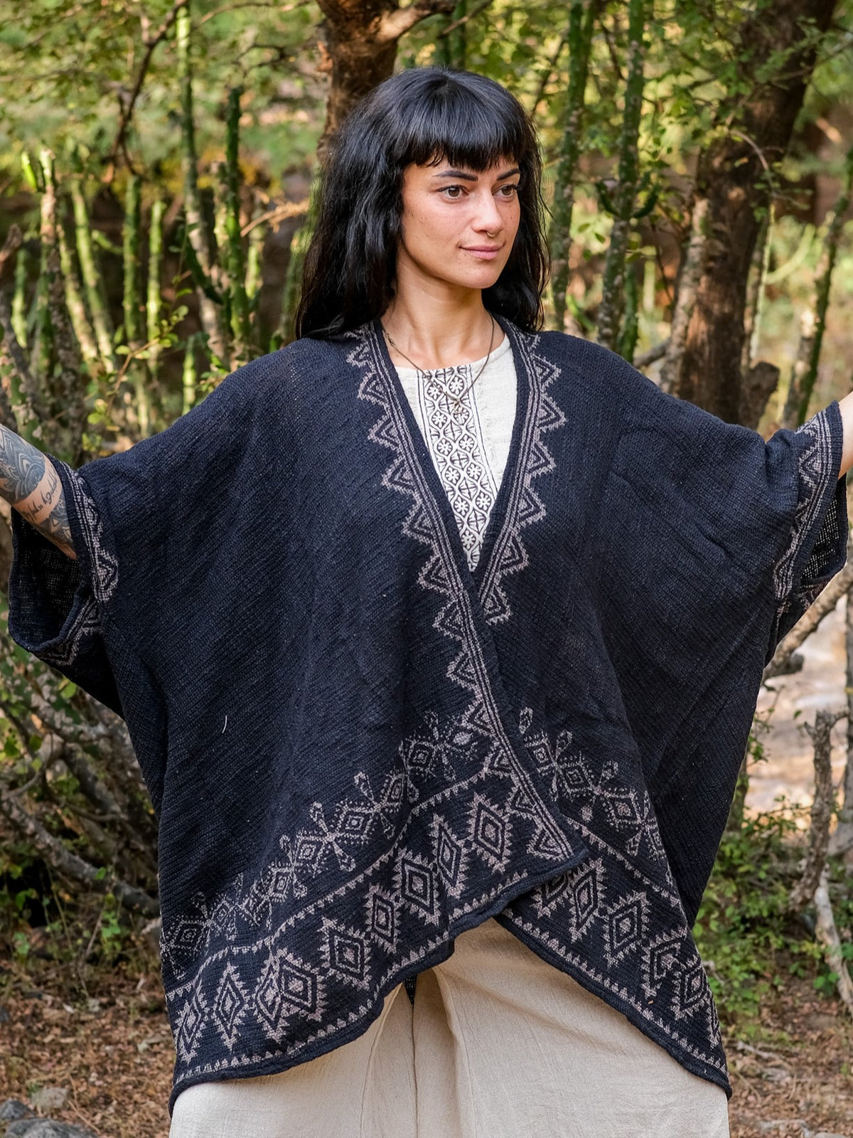 A person stands outdoors with arms slightly raised, wearing the Miraya Shrug in black over a cream outfit. Trees and greenery are visible in the background, highlighting this stylish accessory.