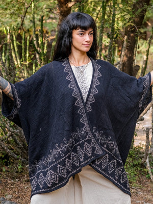 A person stands outdoors with arms slightly raised, wearing the Miraya Shrug in black over a cream outfit. Trees and greenery are visible in the background, highlighting this stylish accessory.