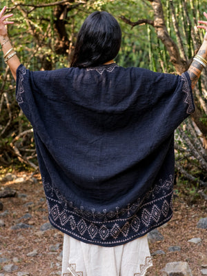 A person with long dark hair, seen from behind, stands outdoors with arms raised. They wear the elegant Miraya Shrug over white clothing, the loose and patterned garment flowing gracefully as they are surrounded by trees and natural scenery.