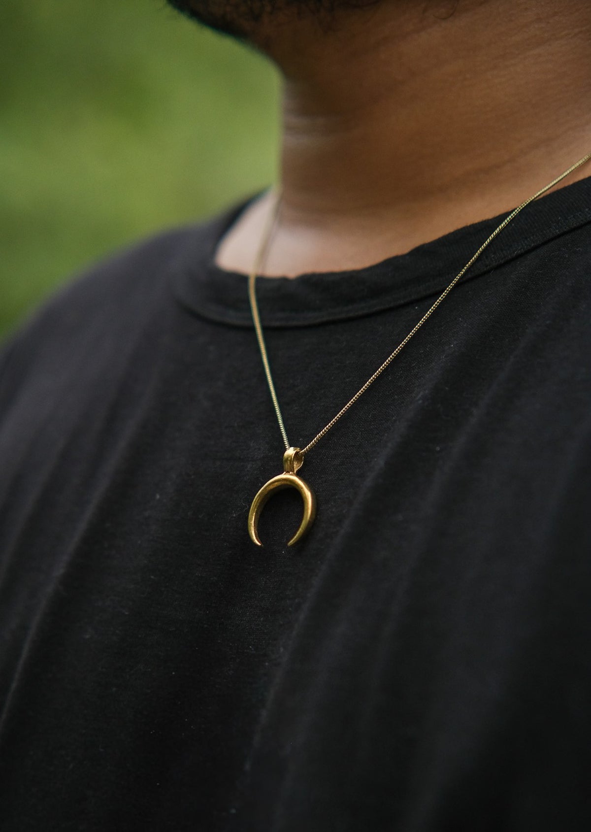 A person wearing the Chandra Talisman, a gold crescent moon pendant necklace, over a black shirt is shown from shoulders to chest with a blurred green background.