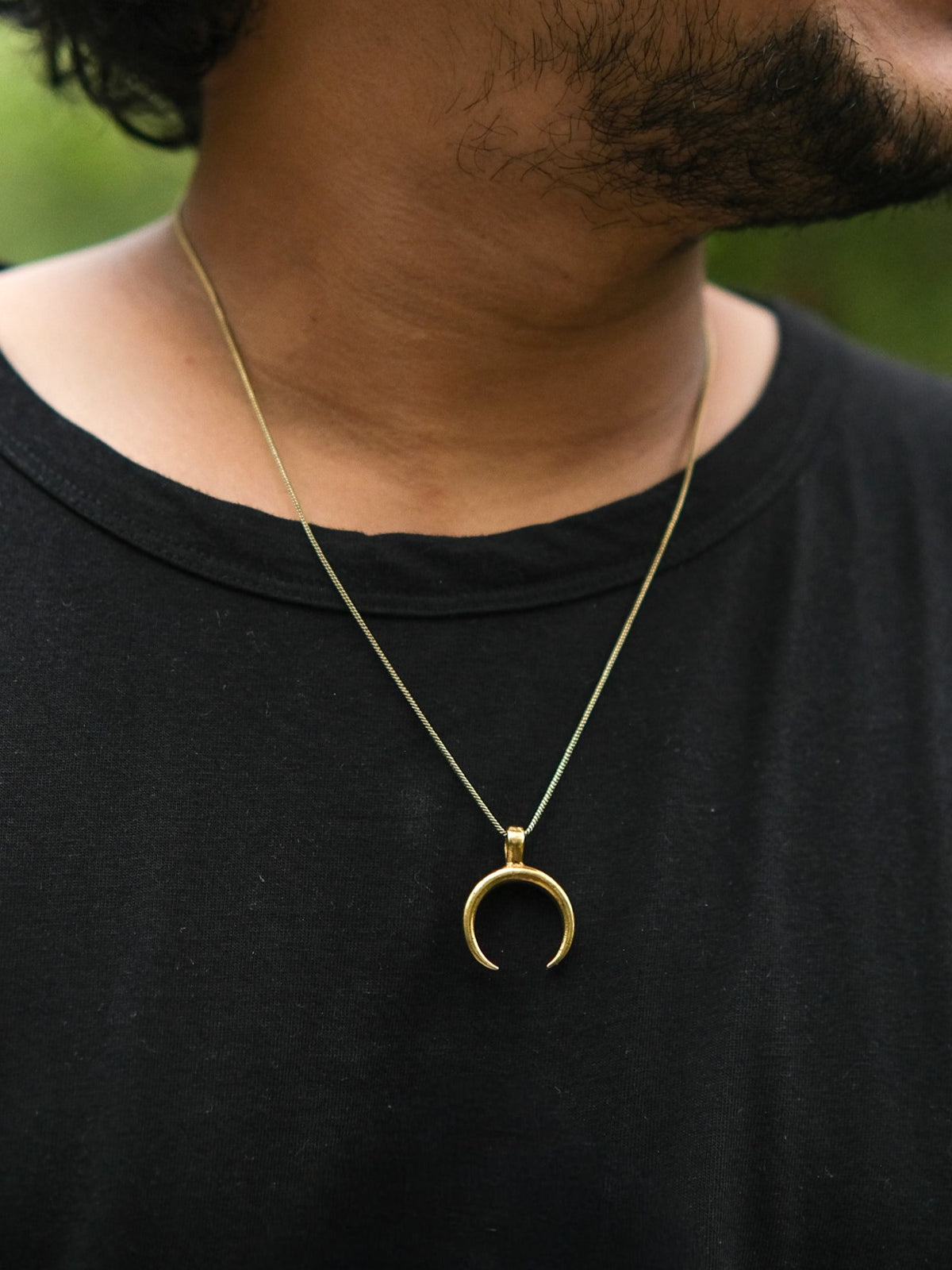 A person wearing the Chandra Talisman gold lunar necklace and a black shirt; only the lower half of their face and neck are visible against a blurred green background.