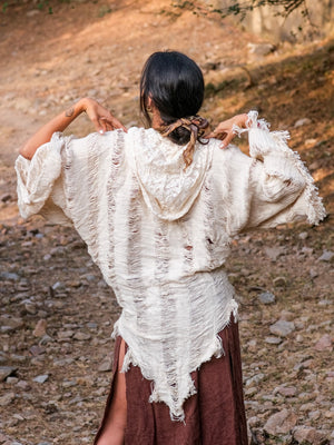 Outdoors on rocky ground, a person with dark hair tied back wears an off-white cotton Kali Shrug with frayed edges over a brown skirt, facing away and raising their hands near their shoulders.