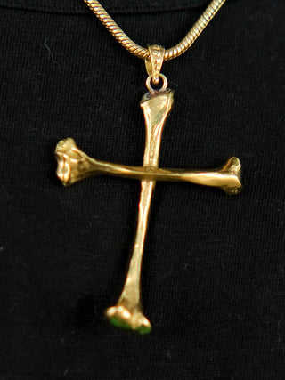 The Eternal Relic Necklace in gold features a pendant of two bone-like pieces, displayed on black fabric—a striking choice for gothic jewelry enthusiasts.