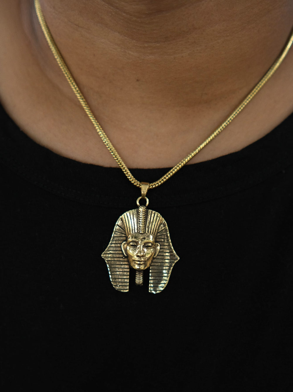 A person dressed in a black shirt wears the Pharaoh Necklace, featuring a gold pendant, adding a touch of classic Egyptian jewelry to their look.