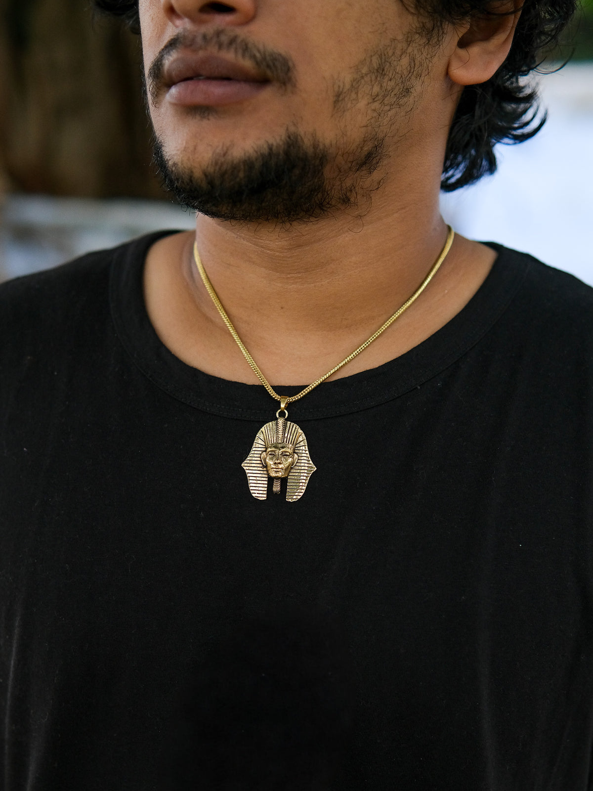 Wearing a black shirt, the person displays their unique taste in Egyptian jewelry with the Pharaoh Necklace, featuring a gold pendant shaped like an ancient Egyptian pharaoh’s mask.