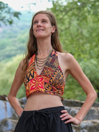 A woman with long brown hair stands outdoors, smiling and looking up. She wears the Tribal Bloom Top, black pants, and a multi-strand necklace. Green trees and rocks are visible in the background.