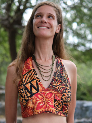A smiling woman with long light brown hair wears the Tribal Bloom Top, featuring colorful geometric patterns and a layered necklace, standing outdoors with blurred green trees in the background.