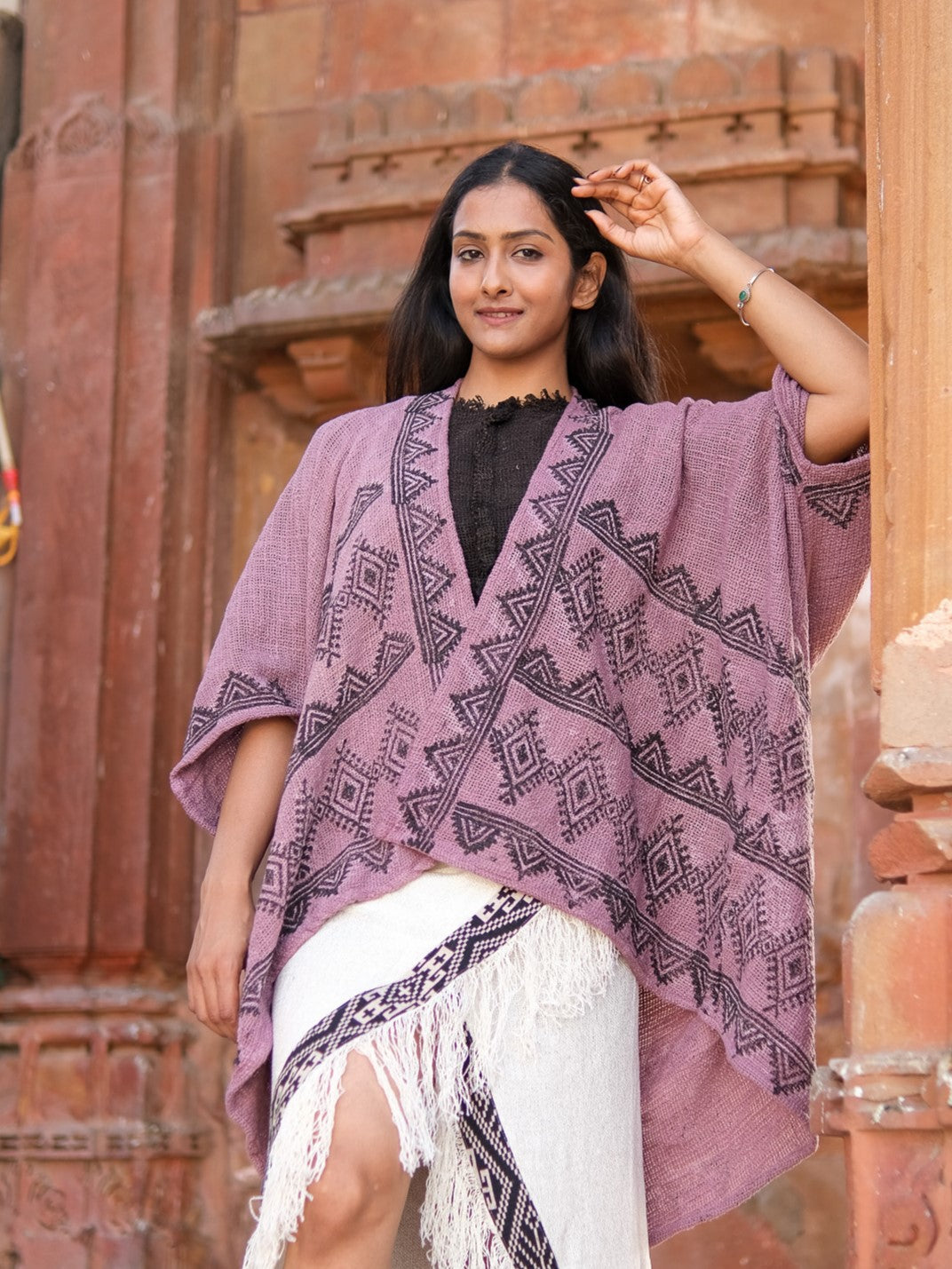 A woman stands outside a reddish-brown stone building, smiling and raising a hand. She wears the Cocoon Shrug—a lavender, hand block-printed cotton piece with black geometric patterns—over a black blouse and white fringed skirt.