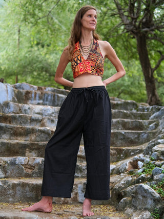 A woman stands barefoot on stone steps outdoors, wearing loose black pants and the Tribal Bloom Top, hands on her hips with green trees behind her—a perfect summer festival look.