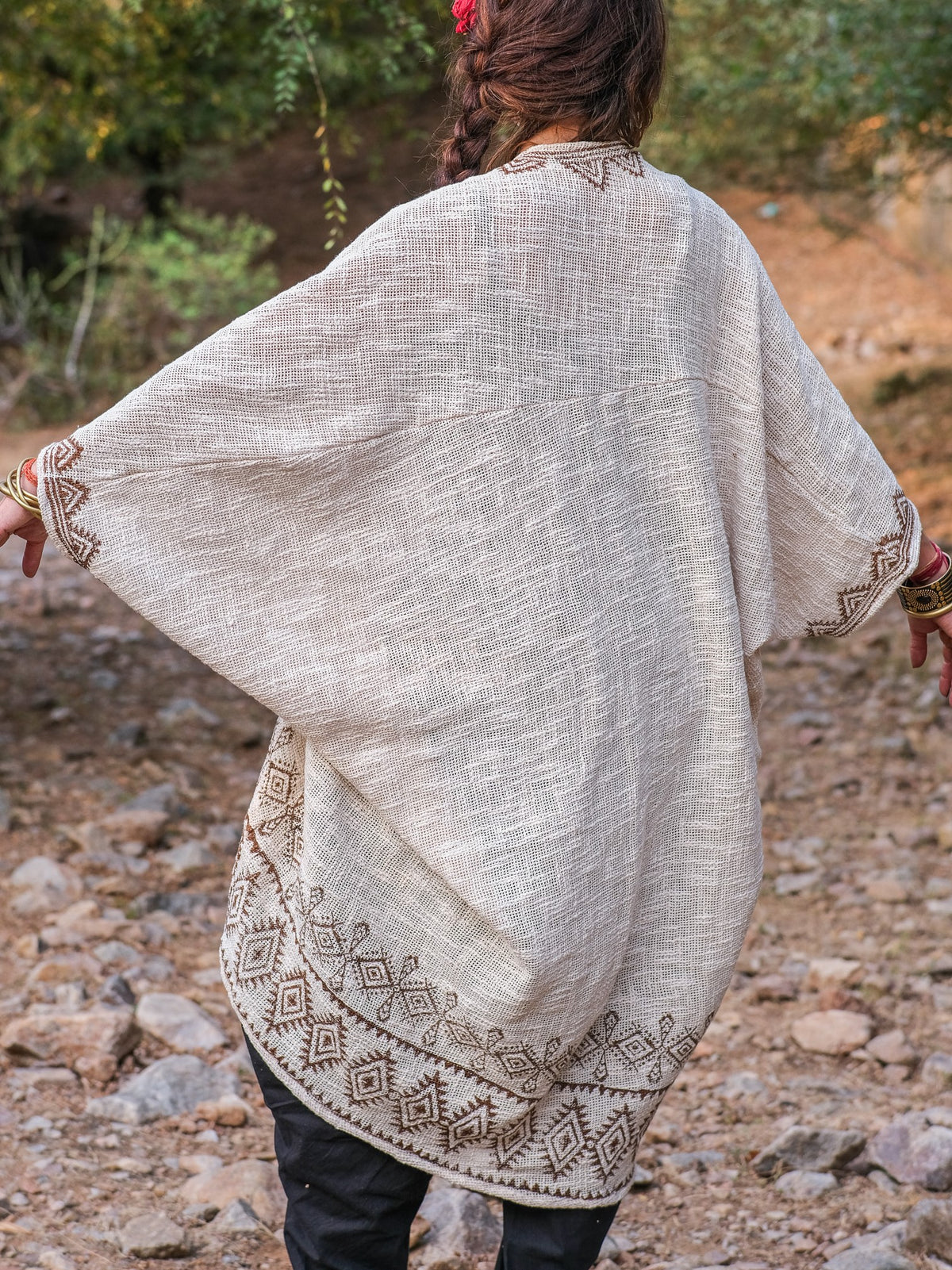 A person stands outdoors with their back to the camera, wearing a cream Miraya Shrug accented by brown geometric embroidery. Their dark hair contrasts with the natural rocks and greenery, highlighting this stylish accessory.