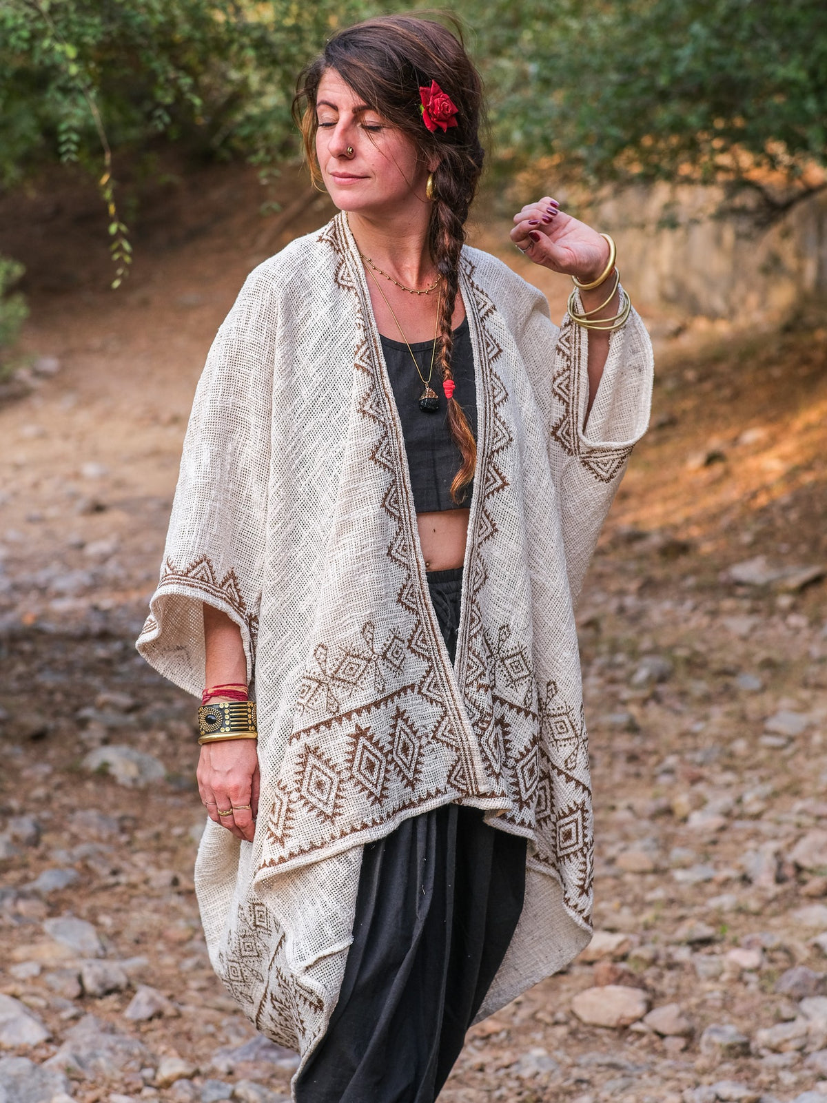 A woman stands outdoors amid trees and rocky ground, wearing a patterned beige Miraya Shrug over dark clothing. A red flower adorns her braided hair, gold bangles grace her wrist, and she exudes a peaceful presence.