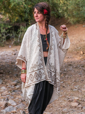 A woman stands outdoors on rocky ground with green foliage in the background, wearing the Miraya Shrug—a stylish, beige-patterned piece—over a black top and black pants. Her braided hair is adorned with a red flower, and she accessorizes with gold bangles.