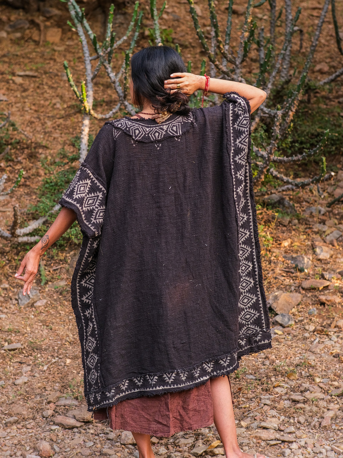 Back view of the model walking away in the black knit kimono with the white block printed geometric border running down the back panels and hem, arms raised, showing the wide sleeves and cobweb trim, on a rocky desert path.