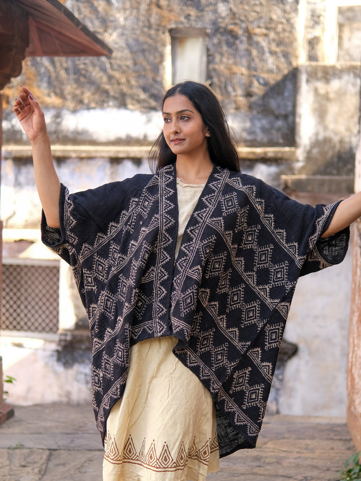 A woman stands outdoors in a hand block-printed, dark blue Cocoon Shrug over a cream dress with geometric designs, arms raised, with a rustic stone building behind her.