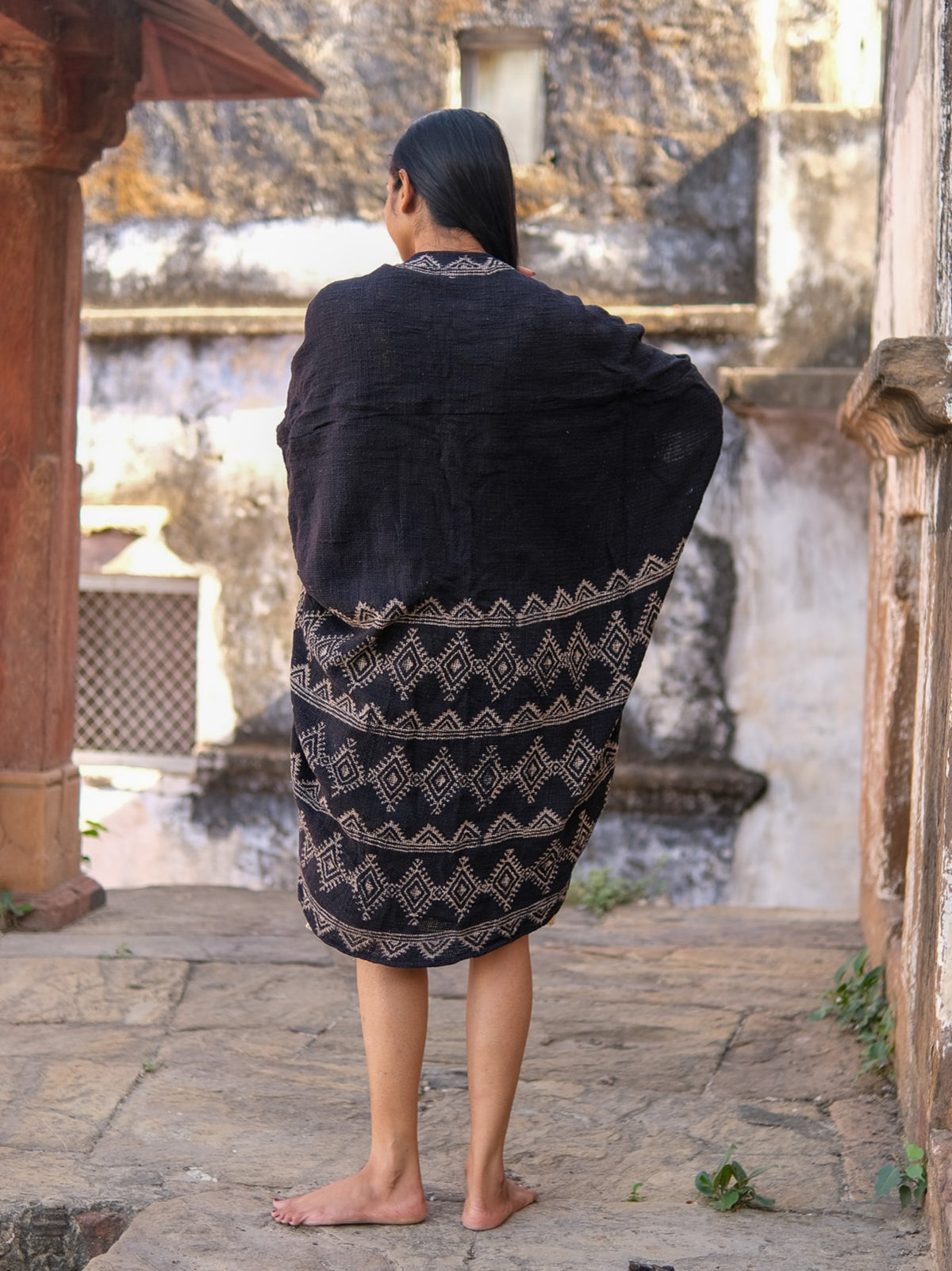 A barefoot person with long dark hair stands on a stone terrace, facing away. They wear the Cocoon Shrug adorned with geometric hand block prints, surrounded by weathered walls and traditional architecture.