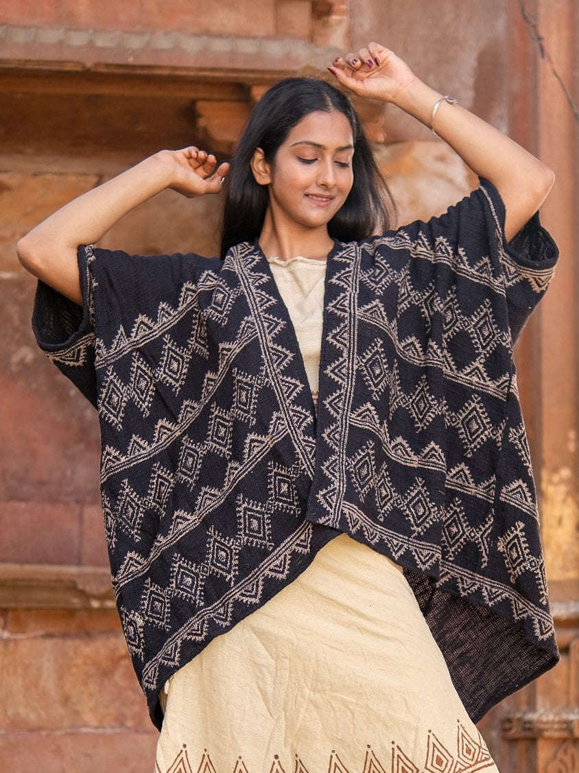 A woman stands smiling with her eyes closed, arms raised, wearing a cream cotton dress and the Cocoon Shrug—a loose black hand block-printed shawl with geometric patterns—in front of a rustic brown wall.