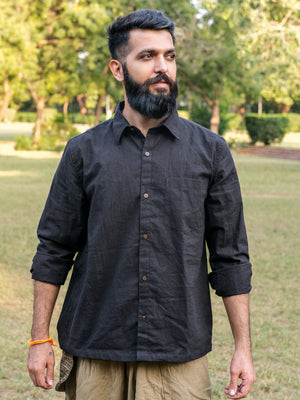 A bearded man stands outdoors on a grassy area, wearing the Bhadra Shirt—a black button-up crafted from premium fabric with rolled-up sleeves—paired with khaki pants. Trees and lush greenery form the background.