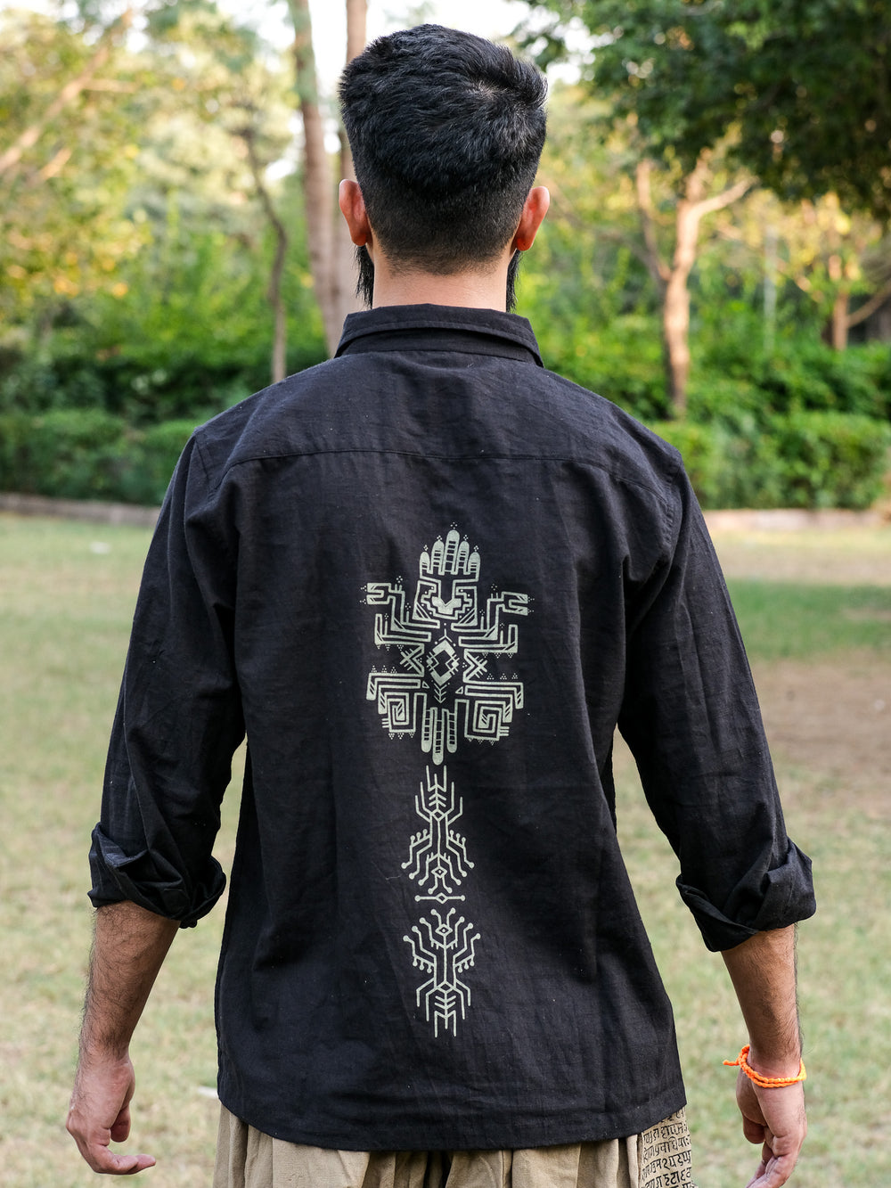 A person stands outdoors facing away, wearing the Bhadra Shirt in black, distinguished by its intricate white geometric patterns on the back. Made from premium fabric, the shirt showcases a unique boho design set against a natural background of trees and grass.