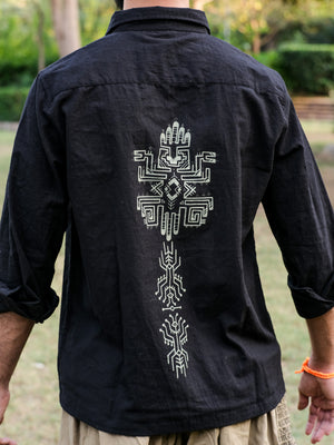 A person wearing the Bhadra Shirt, made from high-quality fabric and featuring detailed white geometric and hand-shaped patterns on the back, is standing outdoors on a grassy area with trees in the background.